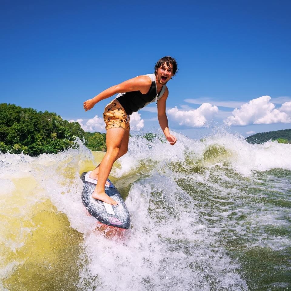 Jordan Koeninger rides a wave on a surfboard, surrounded by water, with green trees and a blue sky with clouds in the background.