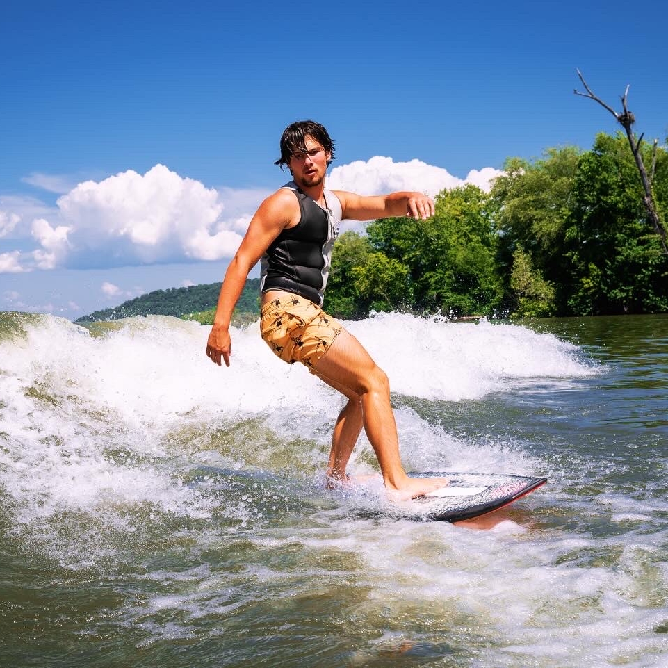 Jordan Koeninger, wearing a life vest and shorts, rides a wakeboard on a river with trees and blue sky in the background.