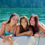 Three young women in swimsuits lean on the edge of a boat, smiling at the camera, with a lake and forested shoreline in the background.