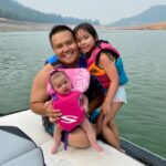 An adult and two children wearing life jackets sit on the edge of a boat, smiling, with a lake and hills in the background.