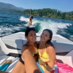 Two women sit on a boat taking a selfie, smiling, while another person wakeboards behind them on a lake surrounded by trees and mountains under a clear blue sky.