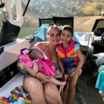 A woman sits on a boat holding a baby in a pink life jacket, with a young child in a colorful life jacket standing beside her. Trees and hills are visible in the background.