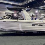 A white Centurion speedboat is displayed indoors at a boat show, with another similar boat visible in the background.