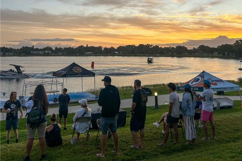 A group of people stand and sit near tents by a lakeside at sunset, with boats on the water and trees in the background.