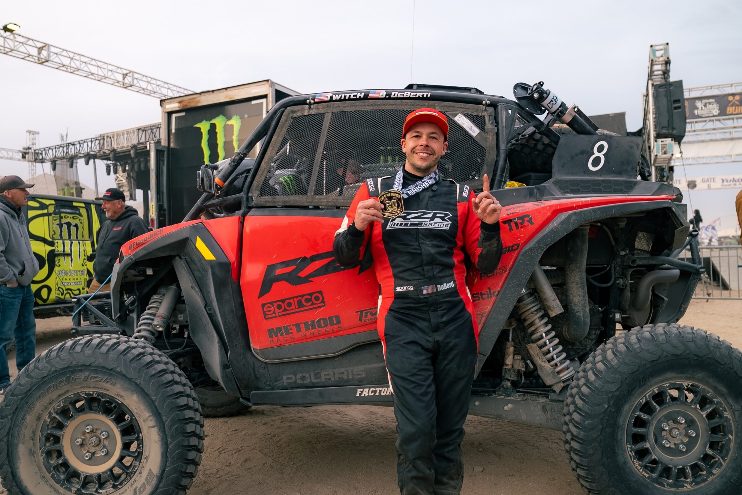 A man in a racing suit stands in front of a red off-road vehicle, holding a medal and smiling at an outdoor event.