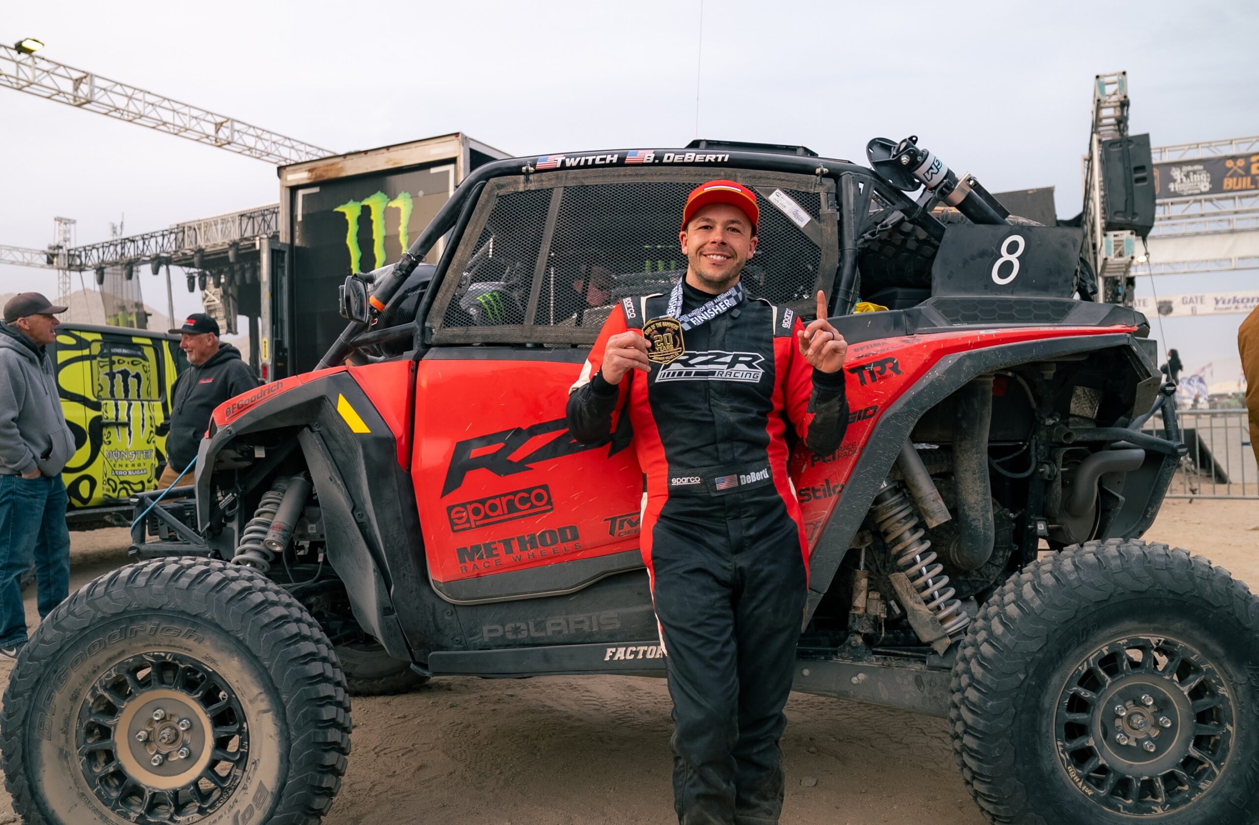 A man in a racing suit stands in front of a red off-road vehicle, holding a medal and smiling at an outdoor event.