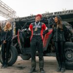 A man in a racing suit with a medal stands smiling in front of an off-road vehicle, surrounded by four women in black outfits on a stage.