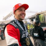 A smiling race car driver in a racing suit and medal stands outdoors at an event, with people and vehicles in the background.