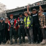 A group of people, including two individuals in racing suits with medals, pose together on a stage under a metal truss at an outdoor event.