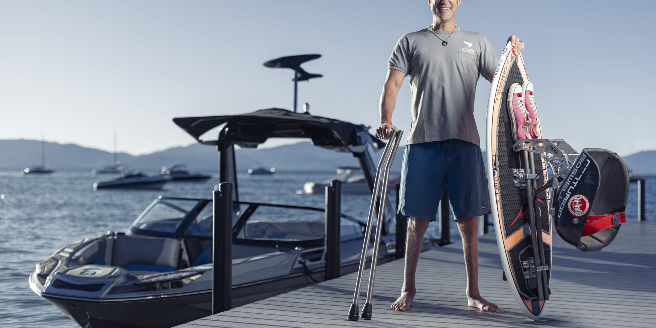 A man with a prosthetic leg stands barefoot on a dock holding a wakeboard and crutches, with a boat and water in the background.