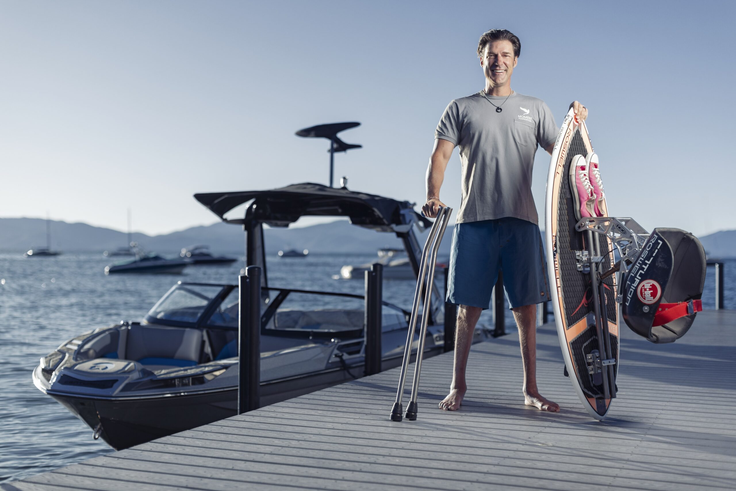 A man with a prosthetic leg stands barefoot on a dock holding a wakeboard and crutches, with a boat and water in the background.