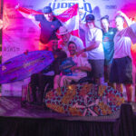 A group of seven people pose on a stage with wakeboards and a trophy in front of a branded event backdrop, under purple lighting.