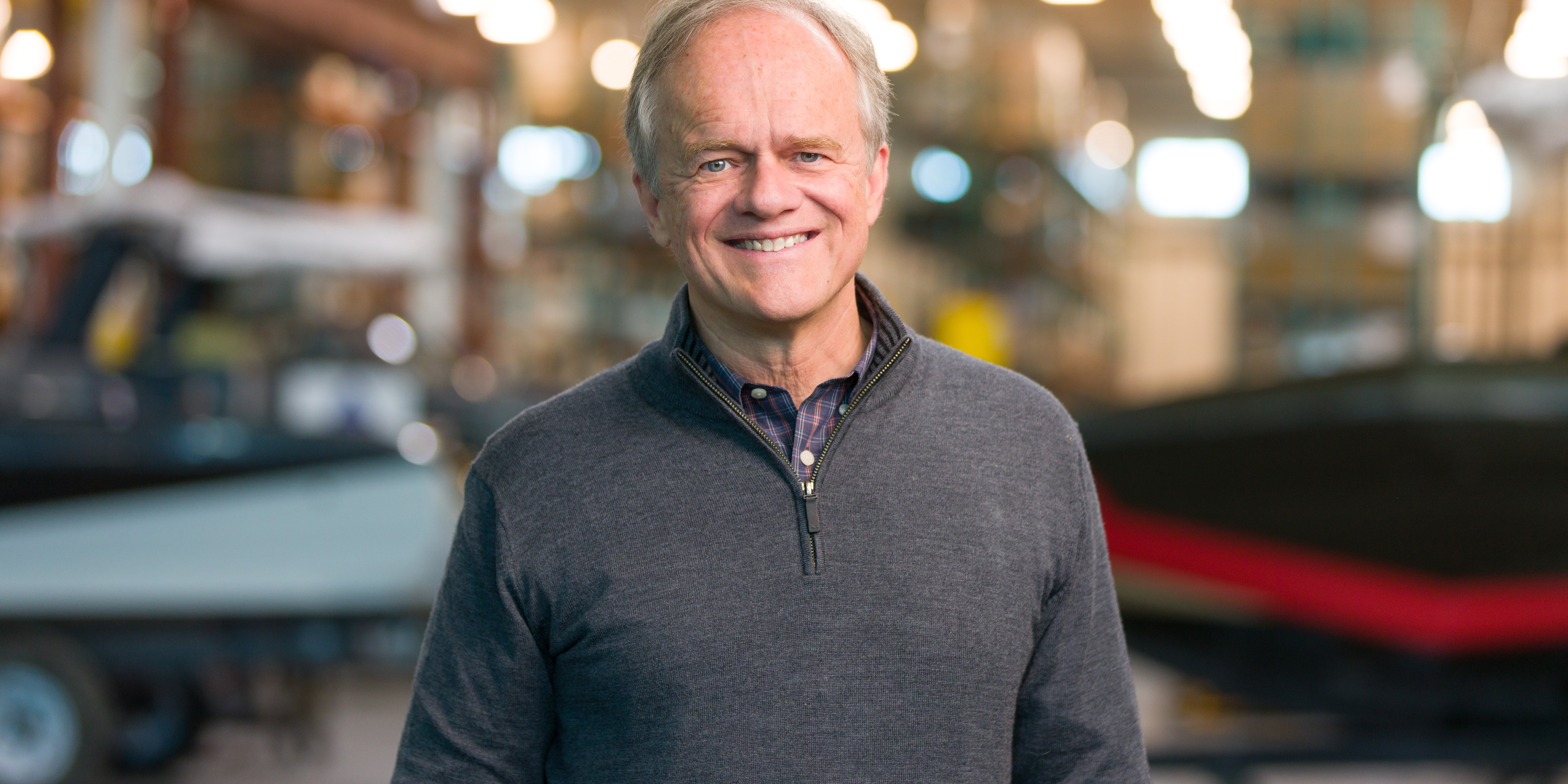 An older man with gray hair smiles while standing indoors, wearing a gray quarter-zip sweater. The background shows blurred boats and equipment in a workshop or warehouse setting.