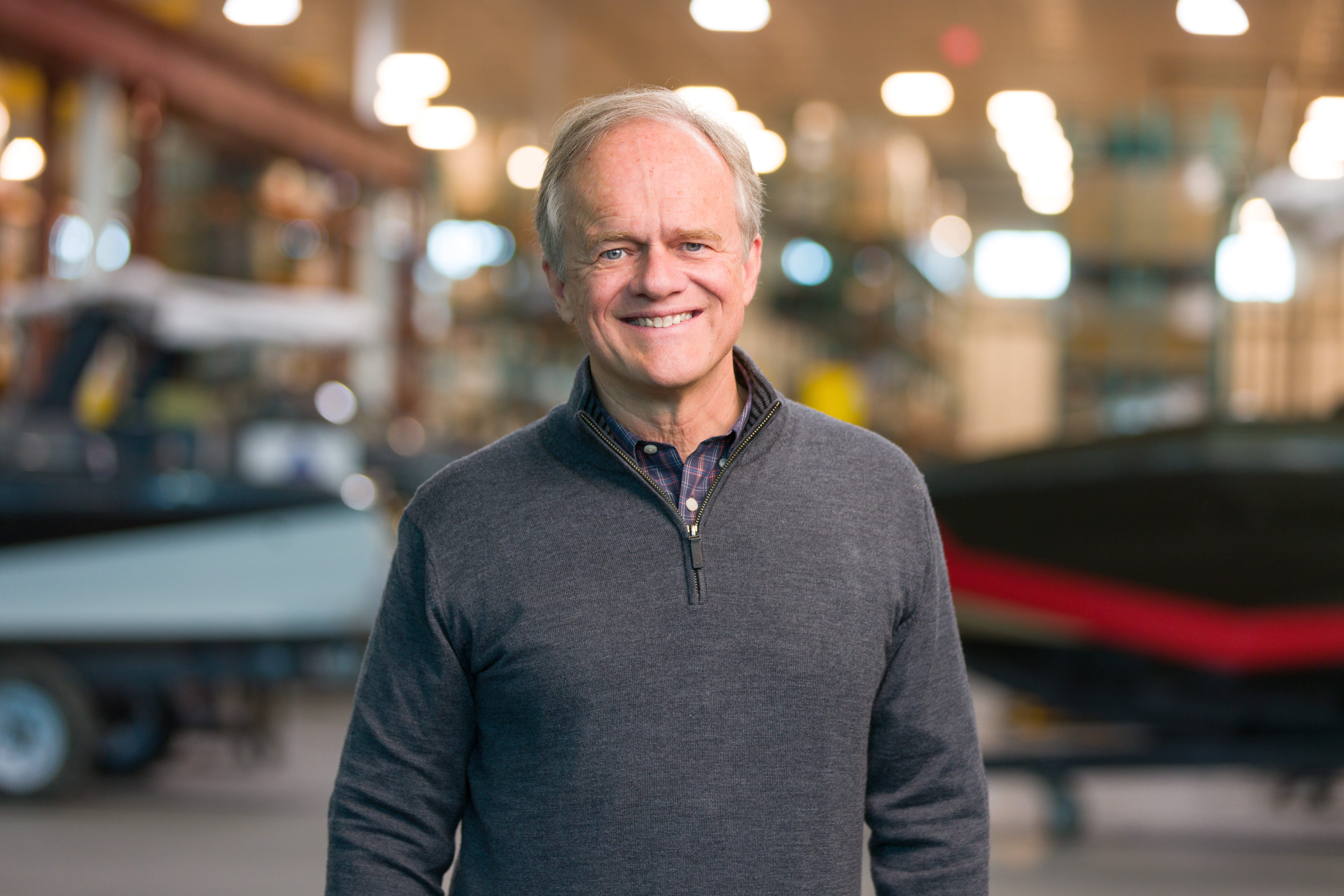 An older man with gray hair smiles while standing indoors, wearing a gray quarter-zip sweater. The background shows blurred boats and equipment in a workshop or warehouse setting.