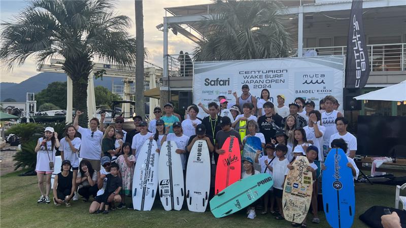 A group of people pose together outdoors in front of a building, holding surfboards at the Centurion Wake Surf Japan Open 2024 event.