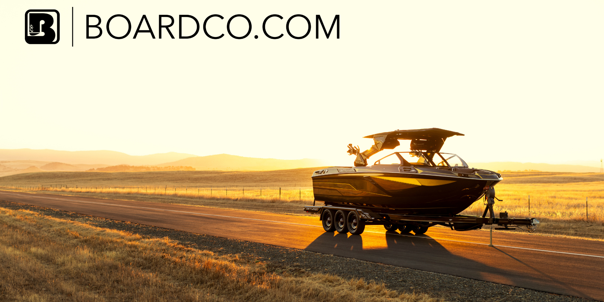 A black boat on a trailer is parked on a rural road at sunset, with fields and hills in the background. The text