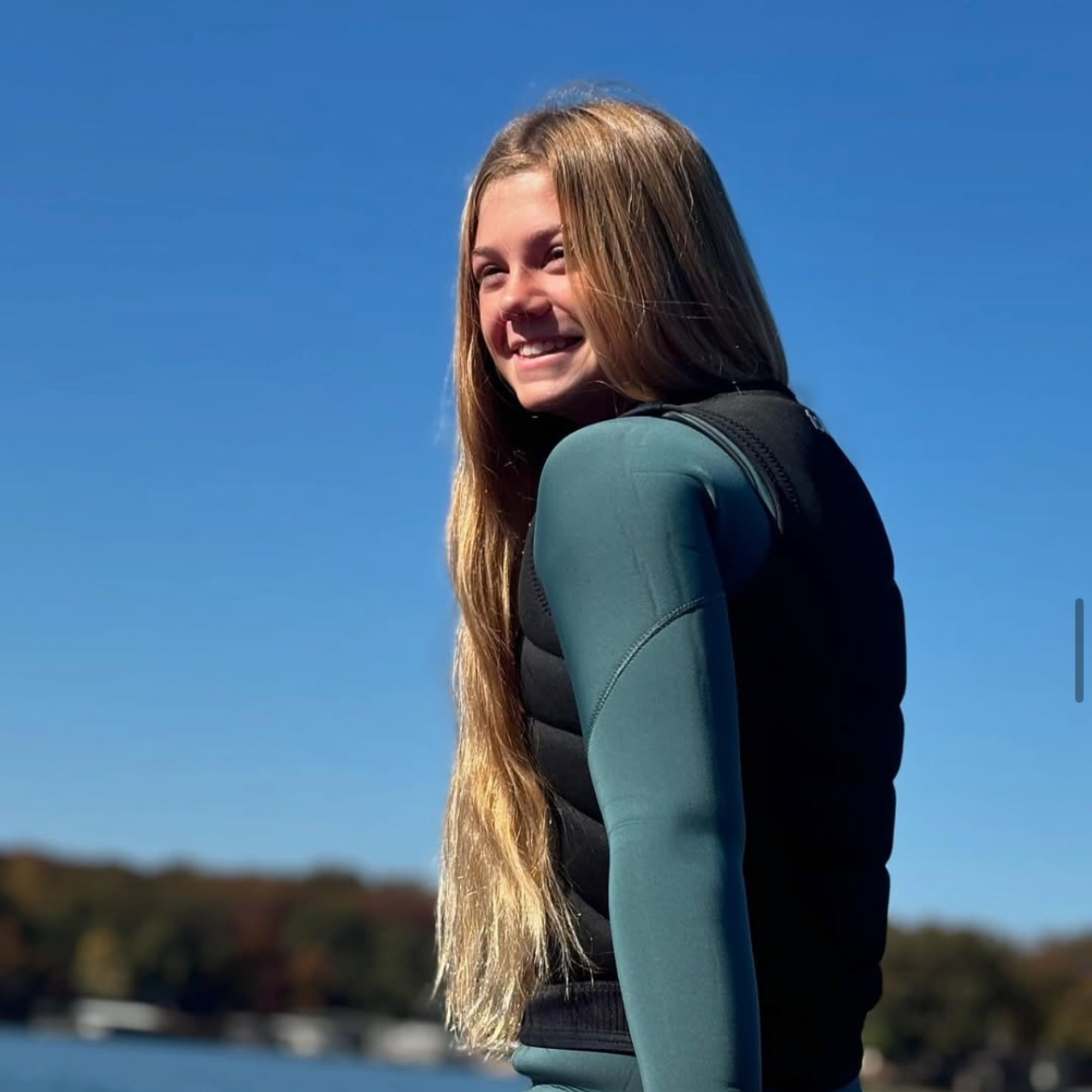 Dylan Ayala, a young person with long blonde hair wearing a wetsuit and life vest, stands by a body of water, smiling under a clear blue sky.