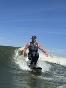 A person wearing a cap, life vest, and wetsuit rides a wave on a surfboard under a clear blue sky.