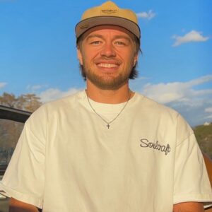 A man wearing a tan cap and white "Soulcraft" shirt stands outdoors, smiling at the camera with blue sky and some clouds in the background.