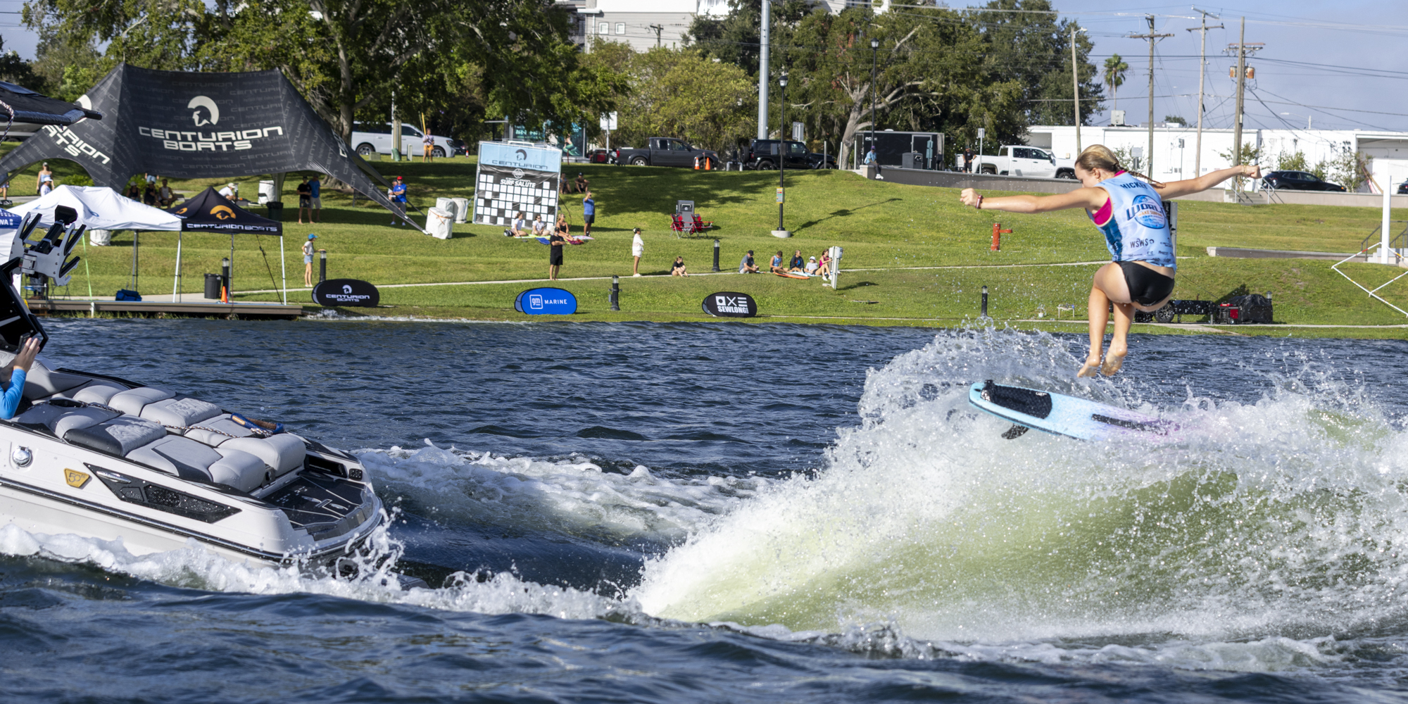 A person wearing a blue vest rides a wakeboard on a wave behind a white boat on a lake, with spectators and event tents on the grassy shore in the background.