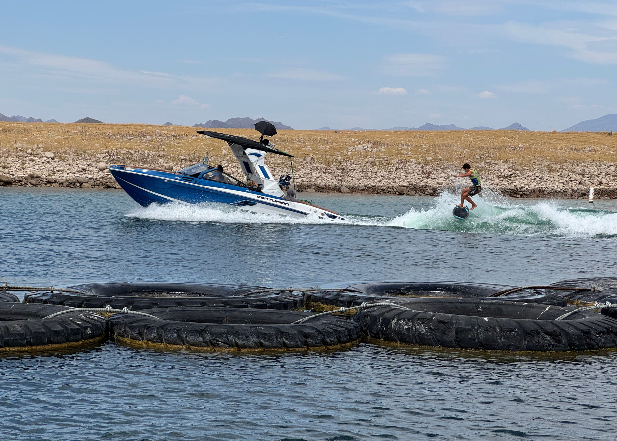 A person wakeboards behind a blue and white motorboat on a lake, with dry, rocky terrain and mountains in the background and large black floats in the foreground.