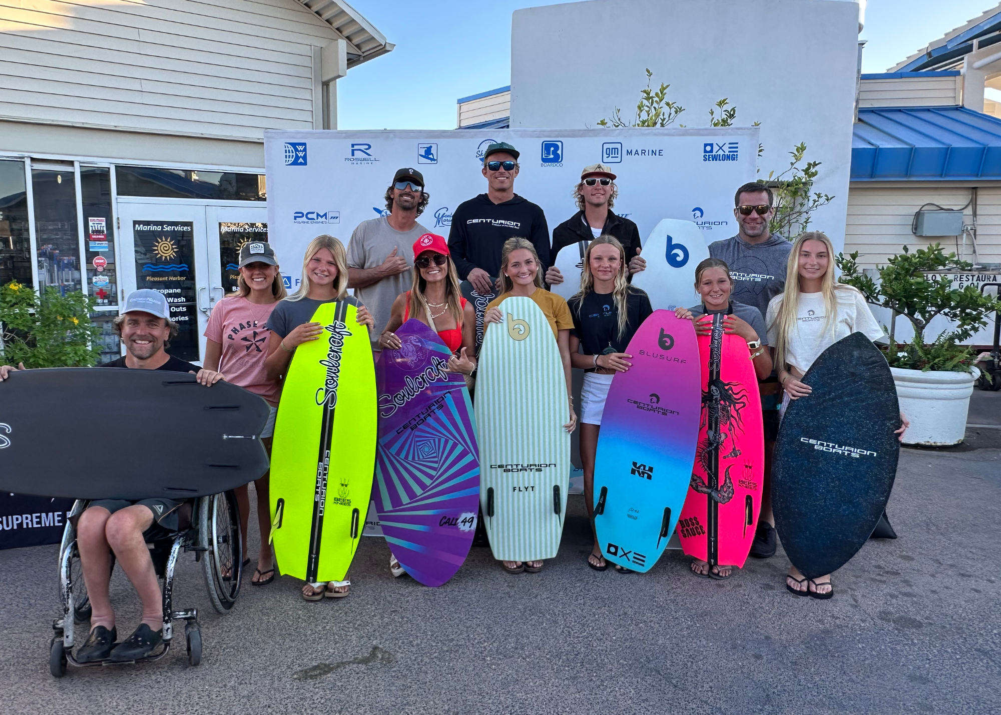 A group of people, some holding colorful bodyboards and surfboards, pose for a photo outside near a white building with a sponsor banner in the background.