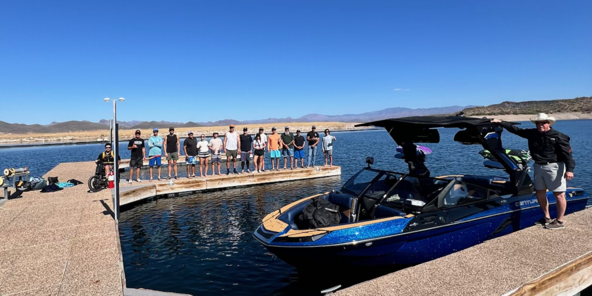 A group of people stand in a line on a dock by the water, with a large blue boat parked nearby under a clear blue sky.