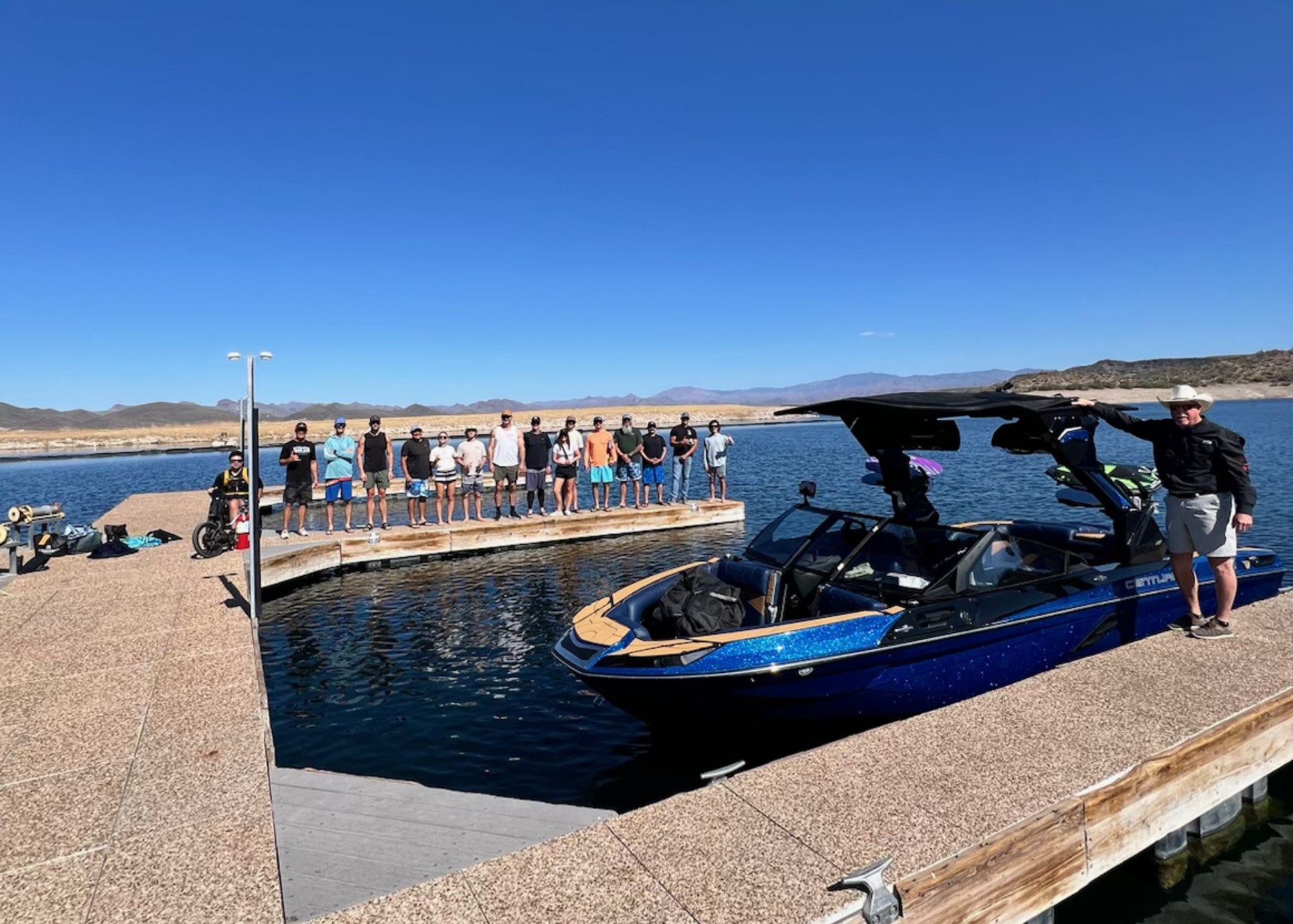 A group of people stand in a line on a dock by the water, with a large blue boat parked nearby under a clear blue sky.