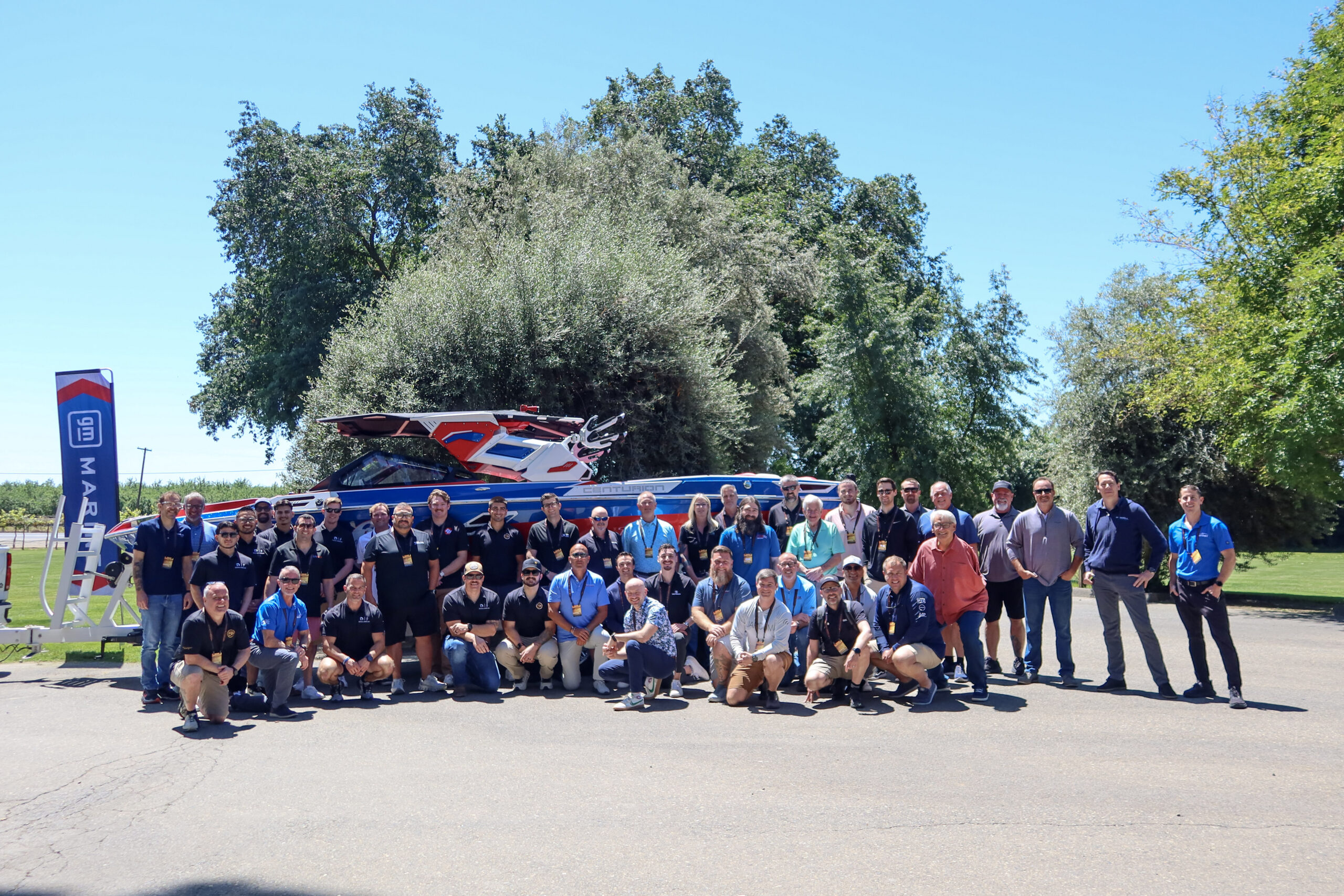 A large group of people pose outdoors in front of a speedboat, with trees and a blue sky in the background.