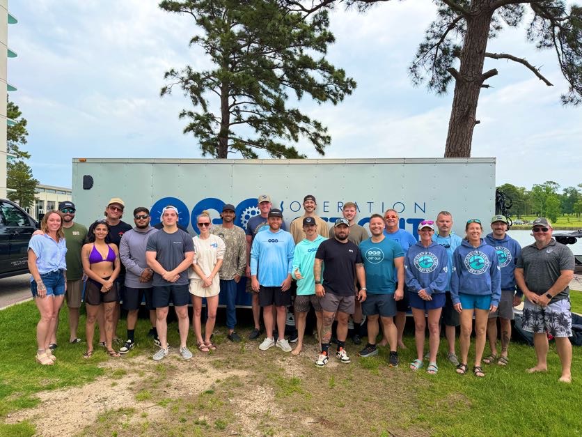 A group of people poses for a photo outdoors in front of a trailer with "Operation" partially visible on the side. Trees and grass are visible in the background.