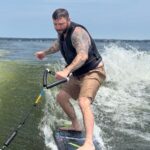 A man with tattoos and a life vest rides a wakeboard on the water, gripping a handle attached to a rope, with waves splashing behind him under a blue sky.