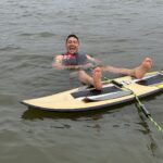 A person in a life jacket floats in the water, smiling and holding onto a paddleboard with their feet up, next to a body of water.