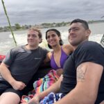 Three people sit closely together on a boat, smiling, with a lake and cloudy sky in the background. The person in the center is wearing a purple swimsuit.