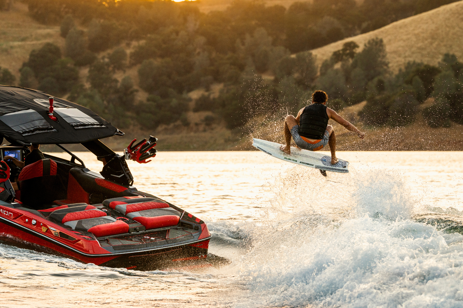 A person on a wakesurf board rides the wake behind a red speedboat on a lake, with hills and trees in the background.