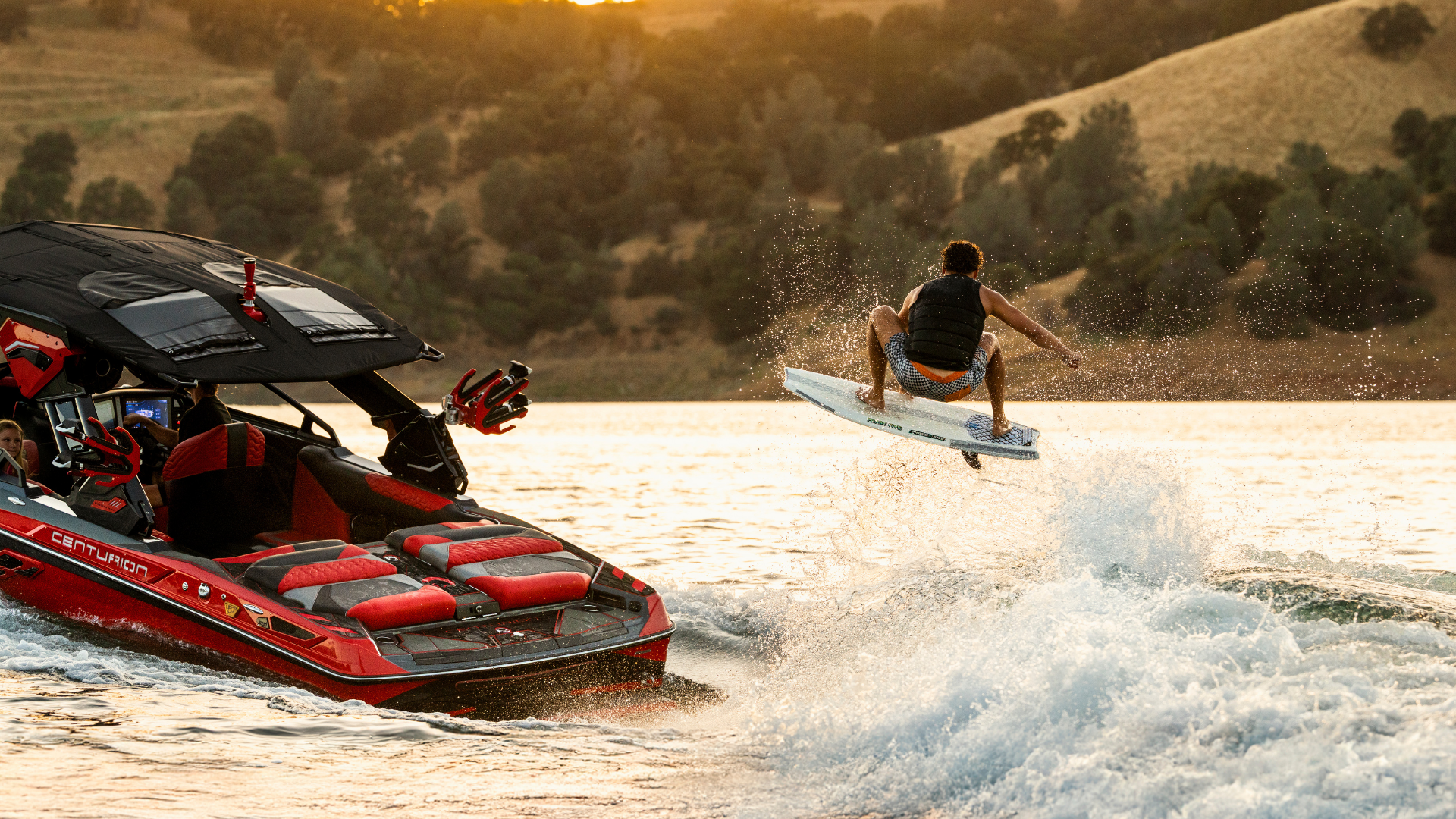 A person on a wakesurf board rides the wake behind a red speedboat on a lake, with hills and trees in the background.