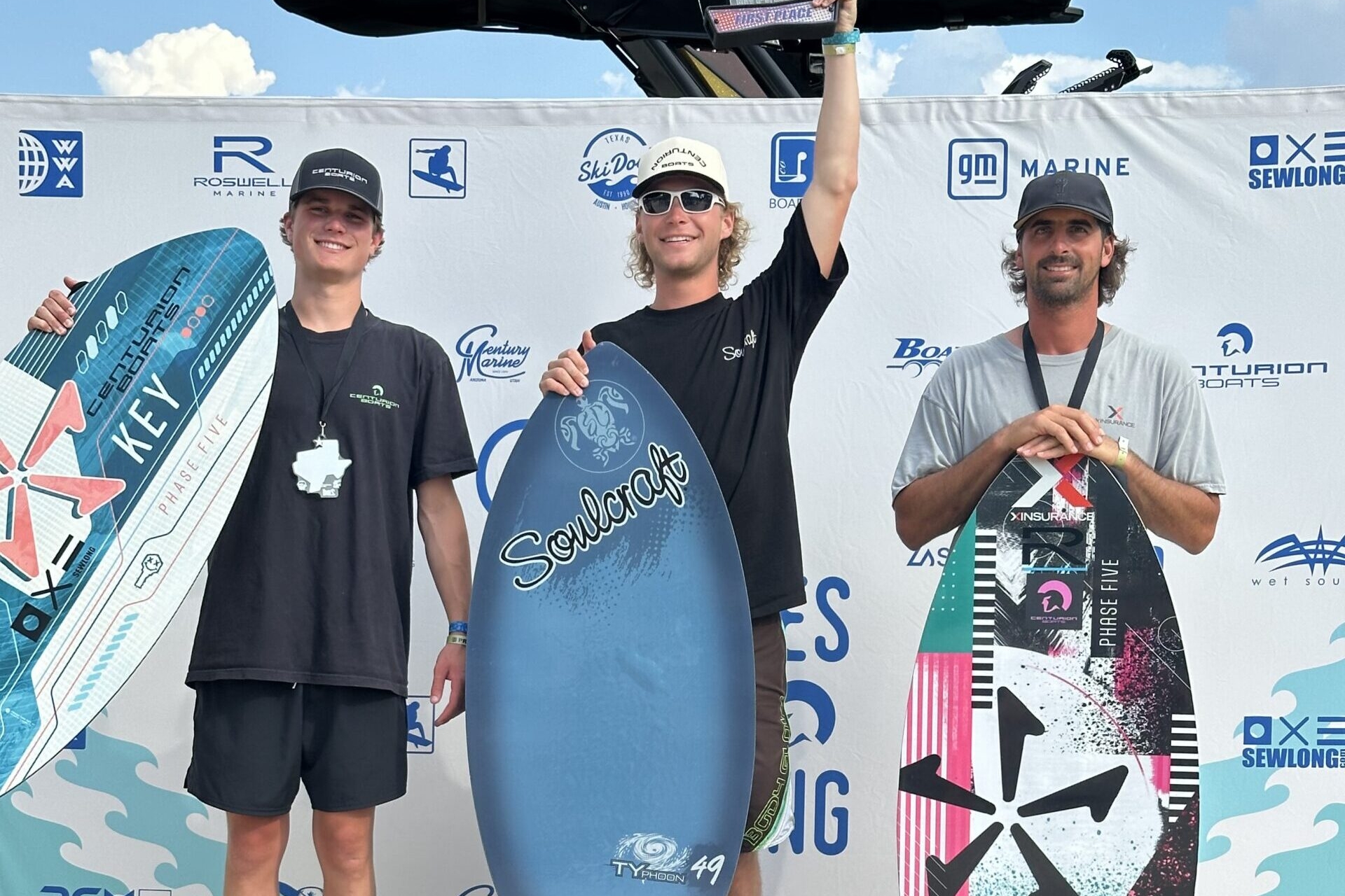 Three men stand on a winners' podium holding wakeboards and trophies at the World Series of Wake Surfing event, with a branded backdrop behind them.