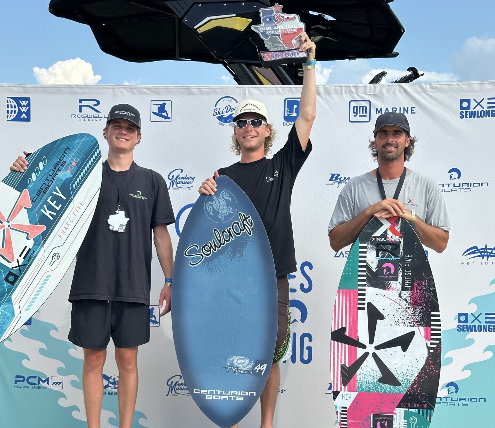 Three men stand on a winners' podium holding wakeboards and trophies at the World Series of Wake Surfing event, with a branded backdrop behind them.