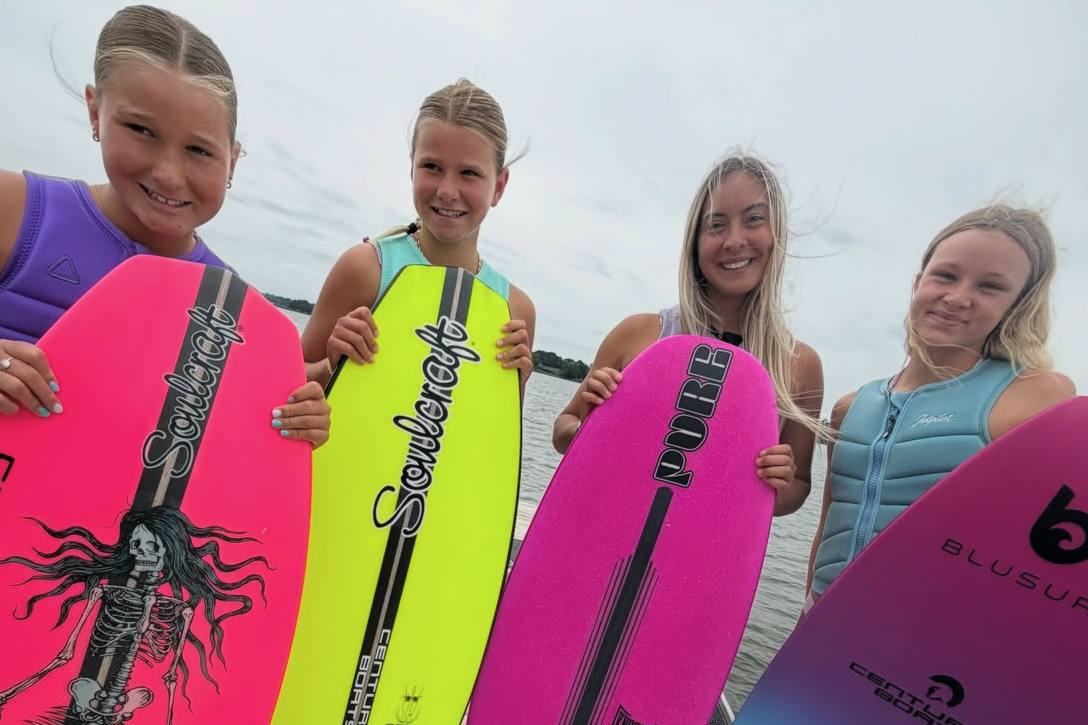 Four young girls stand on a dock holding colorful wakesurf boards, smiling at the camera. The sky is overcast and water is visible in the background.