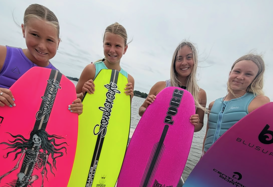 Four young girls stand on a dock holding colorful wakesurf boards, smiling at the camera. The sky is overcast and water is visible in the background.