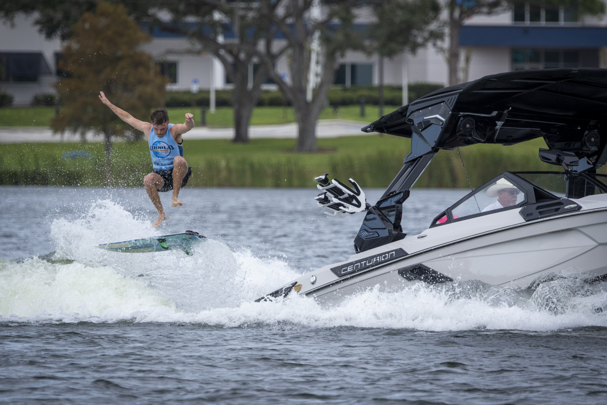 A person in a tank top jumps off a wakeboard behind a moving speedboat on a lake, with trees and buildings in the background.
