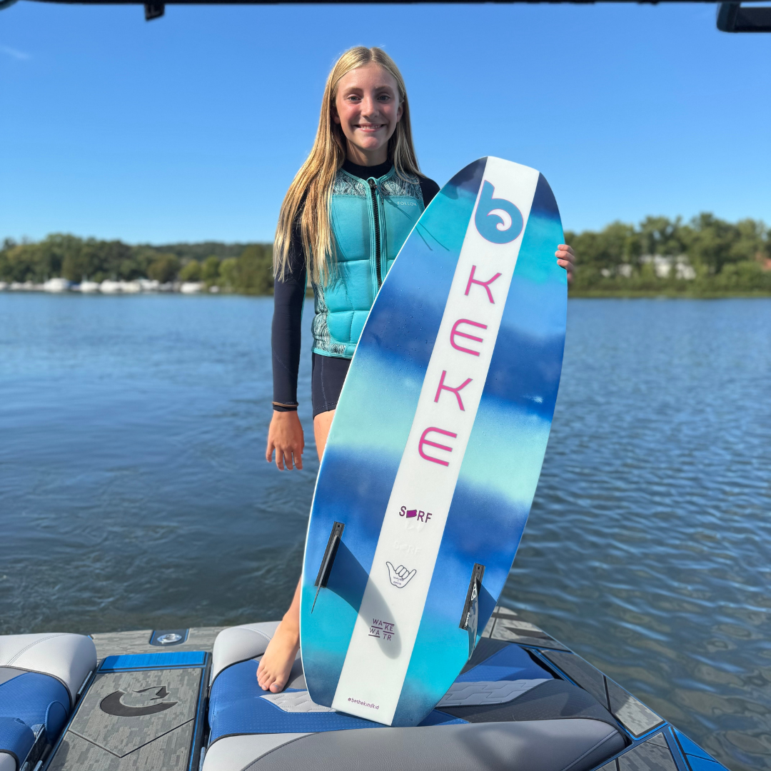 A girl in a blue life vest stands on a boat holding a blue and white surfboard with 