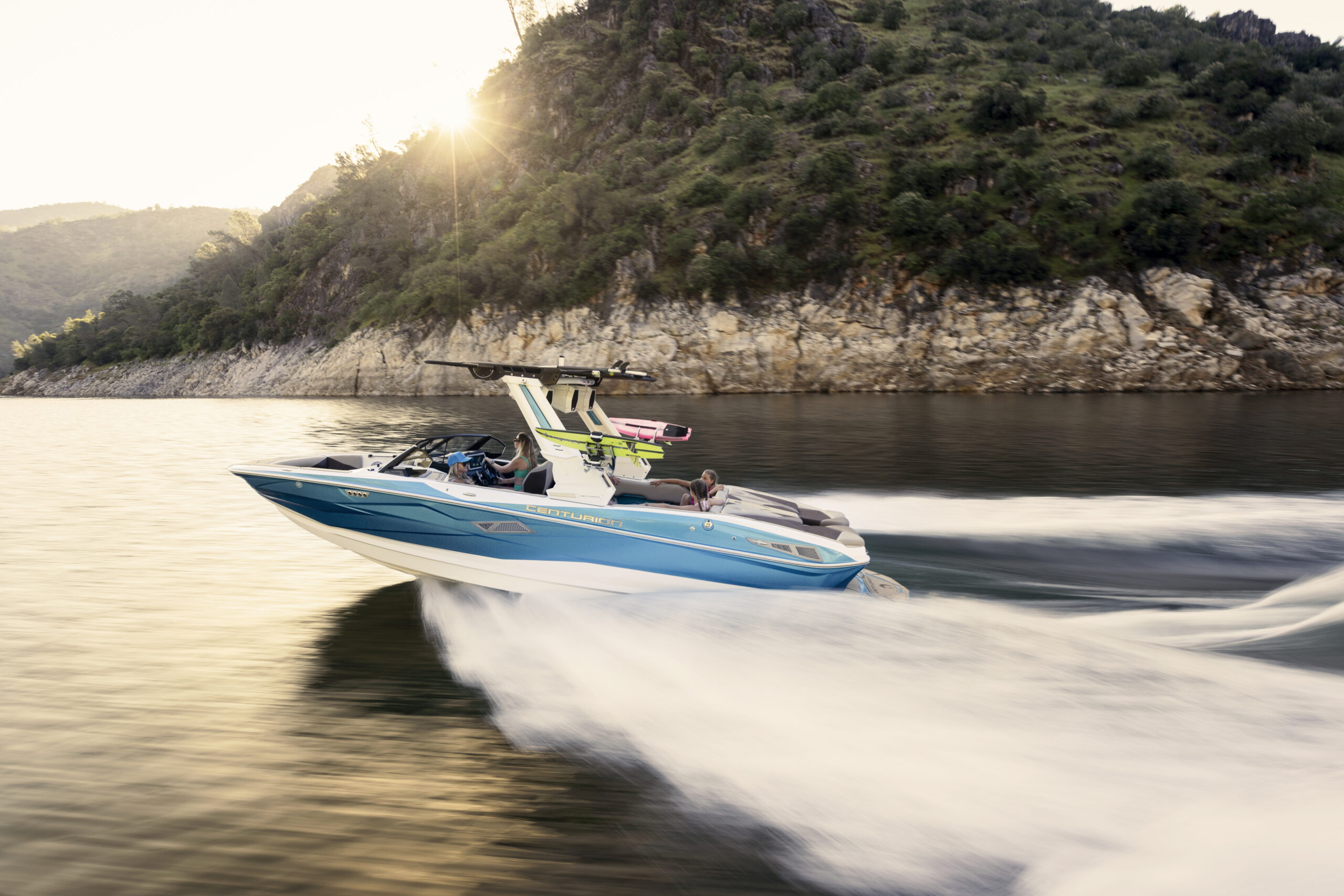 A blue and white speedboat with people onboard moves quickly across a lake, creating waves, with a forested hillside and sunlight in the background.