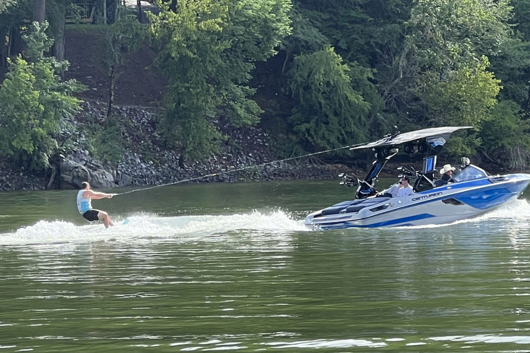 A person is wakeboarding on a river, being towed by a blue and white motorboat with several people onboard. Trees line the riverbank in the background.