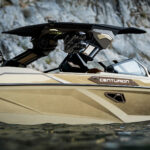 A tan Centurion boat floats on the water near rocky cliffs, showing a close-up of its side, windshield, and wakeboard tower.