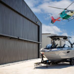 A silver motorboat on a trailer is parked near an airport hangar, with a helicopter flying overhead and an air traffic control tower in the background.