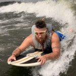 A bearded man wearing sunglasses and a life vest rides a bodyboard on a wave, surrounded by splashing water.