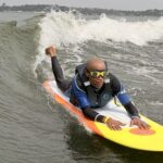 A person in a wetsuit and sunglasses paddles on a yellow surfboard, riding over a small ocean wave with land visible in the background.
