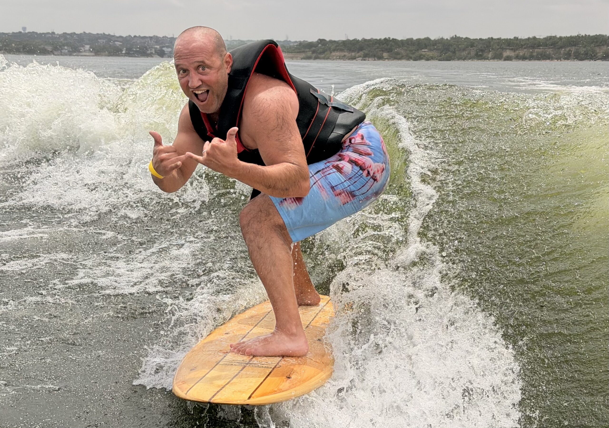 Bald man in a life vest surfs on a wooden board, smiling and making a hand gesture, with water splashing behind him.