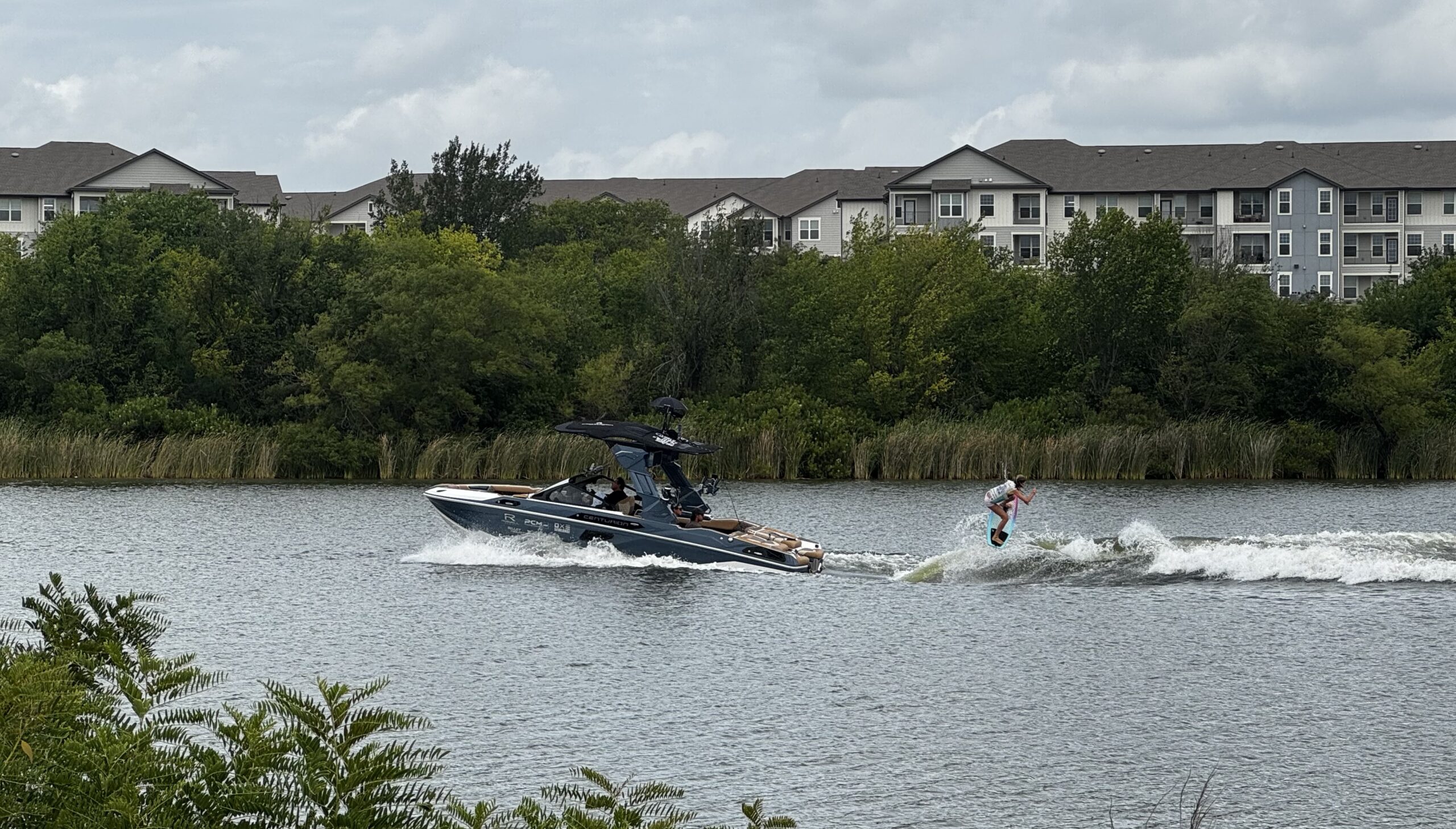 A person wakeboards behind a motorboat on a lake, with trees and apartment buildings visible in the background under a cloudy sky.