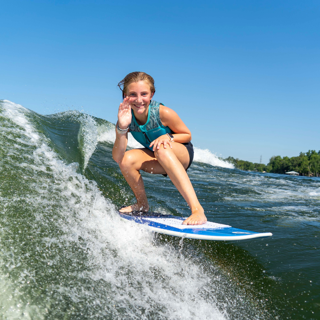 A young woman in a teal vest surfs on a wave, crouching on her board and smiling while waving with her right hand. The sky is clear and blue.